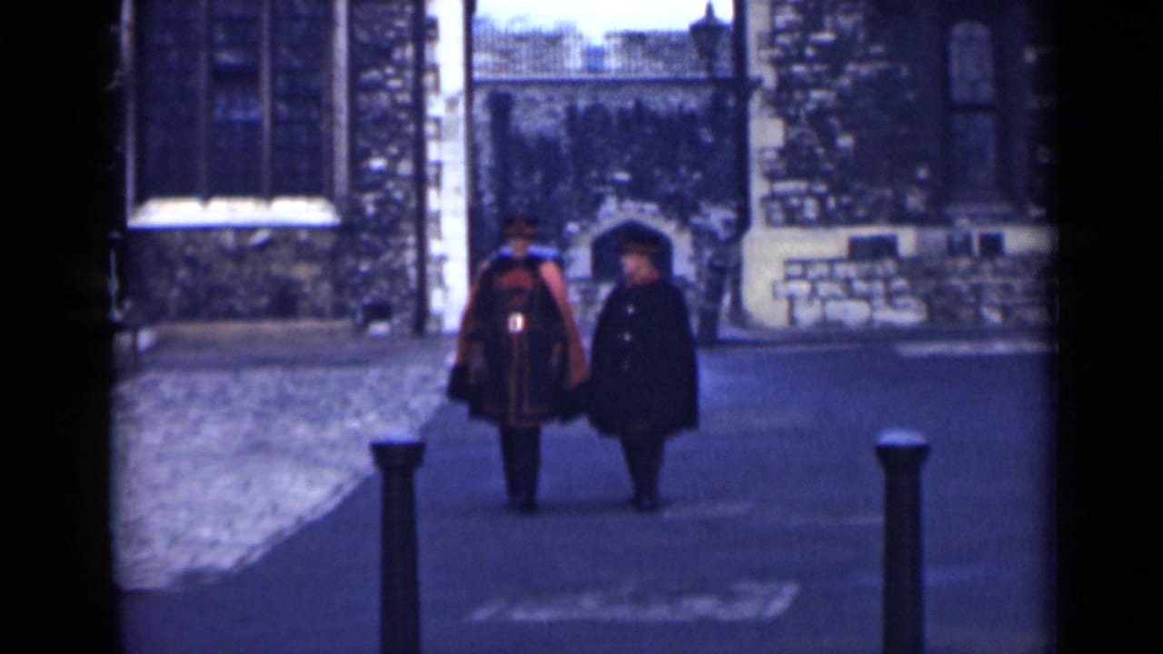 Two men in formal outerwear walk closer to the camera in a London stone courtyard, with a gate and arched doorway behind them.