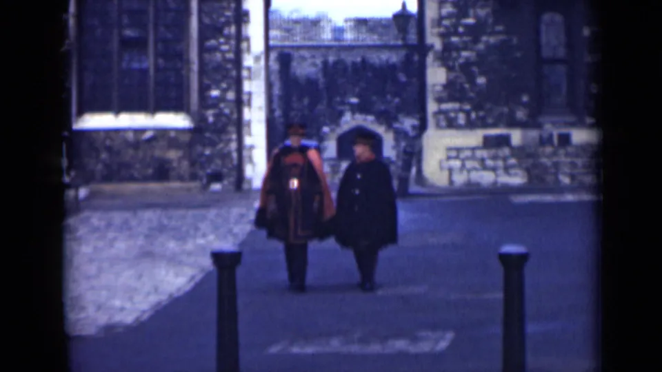 Two men in formal outerwear walk closer to the camera in a London stone courtyard, with a gate and arched doorway behind them.