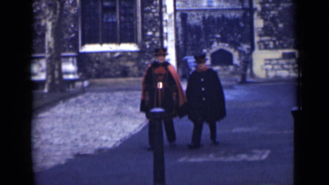 Two men in formal clothing walk along a cobblestone street beside a stone building.