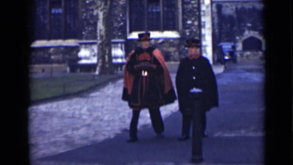 A closer 1946 London frame shows the red-and-black uniform, dark overcoat, hats, cobblestones, and a stone building behind the walking pair.