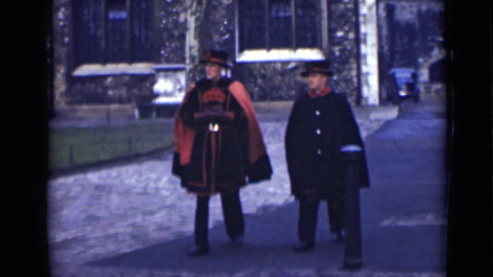 At the end of the short London clip, the uniformed man and companion walk side by side, framed by cobblestones, bollards, and a historic stone exterior.