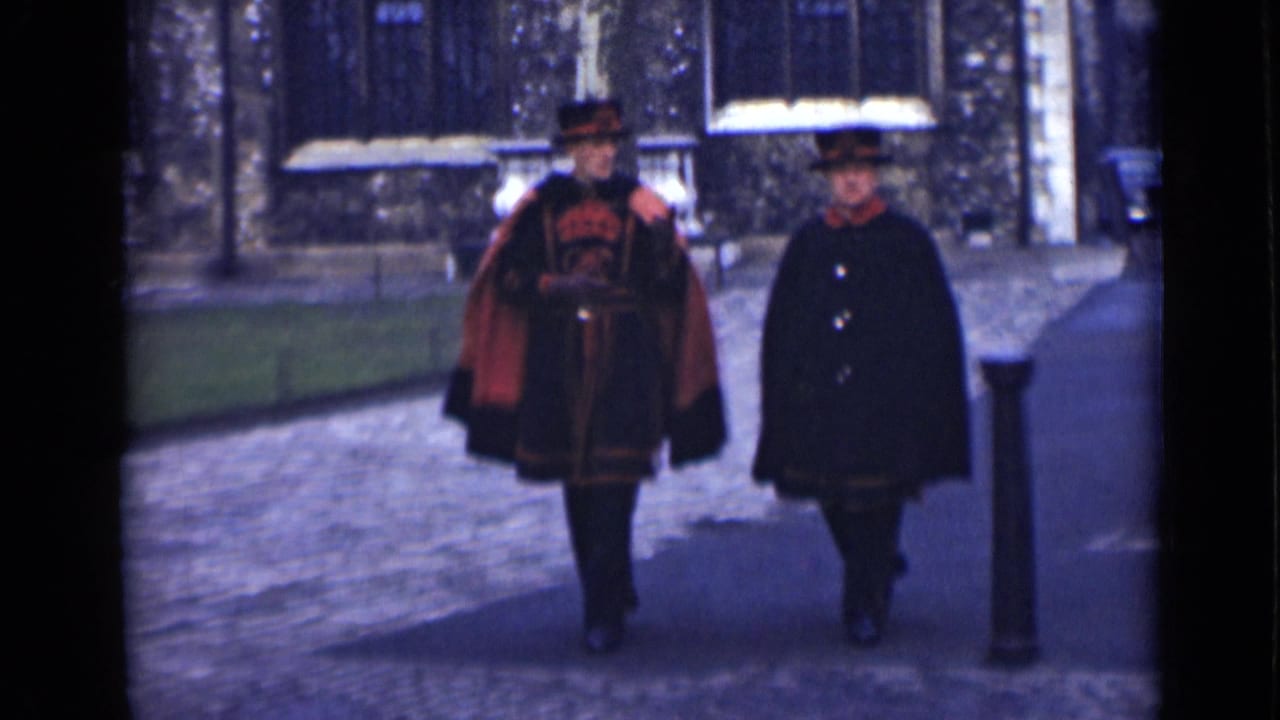 Two adults in dark outerwear walk through a London garden setting in 1946, with stone buildings and grass behind them.