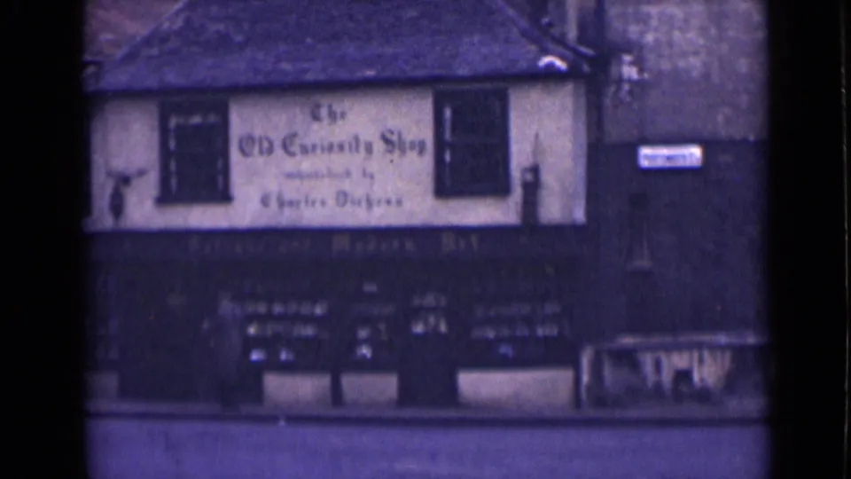A London shopfront appears with a readable Old Curiosity Shop sign, dark lower facade, windows, and street benches in soft 8mm color.