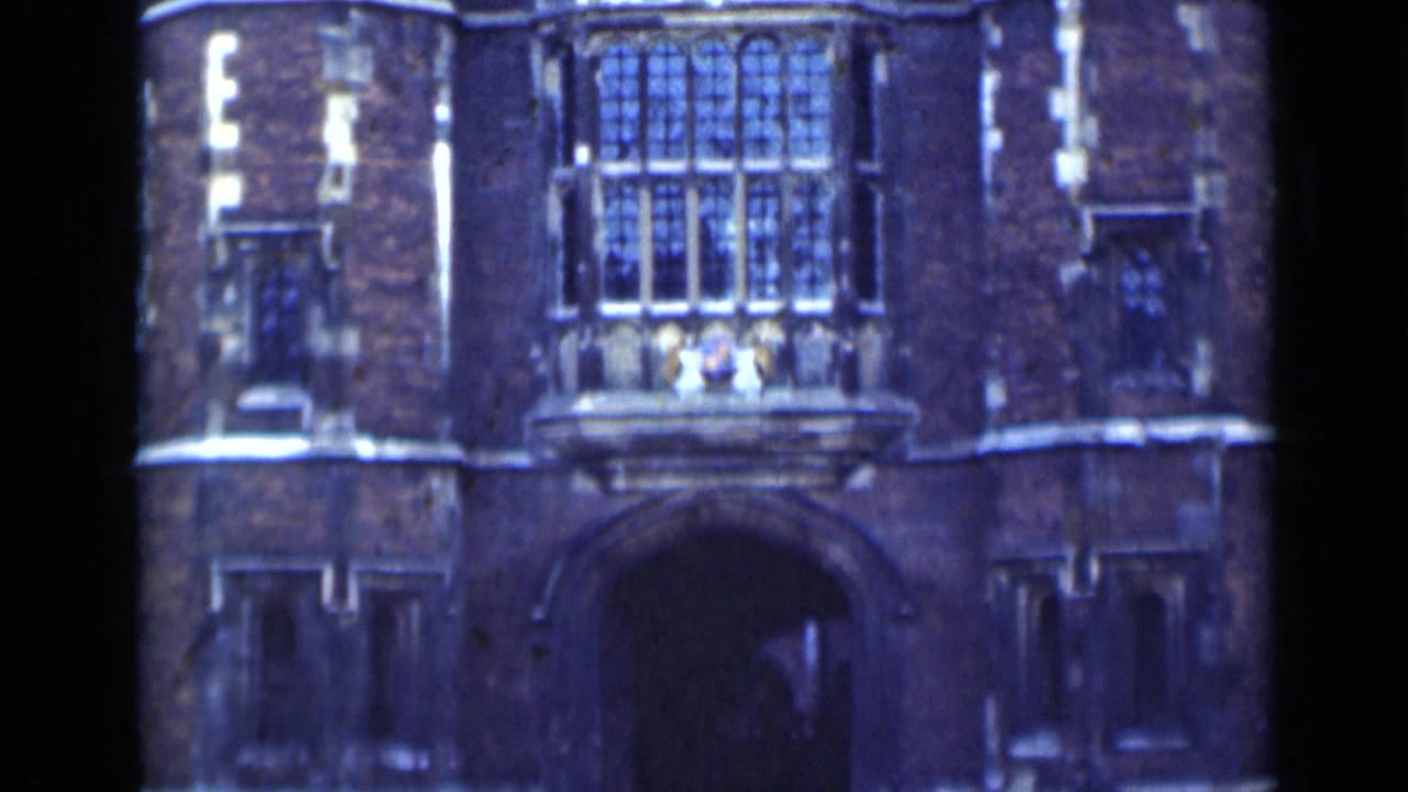 1940s London 8mm frame showing a brick historic building entrance with Gothic-style windows, pale stone trim, and a deep arched doorway.