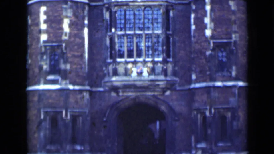 1940s London 8mm frame showing a brick historic building entrance with Gothic-style windows, pale stone trim, and a deep arched doorway.