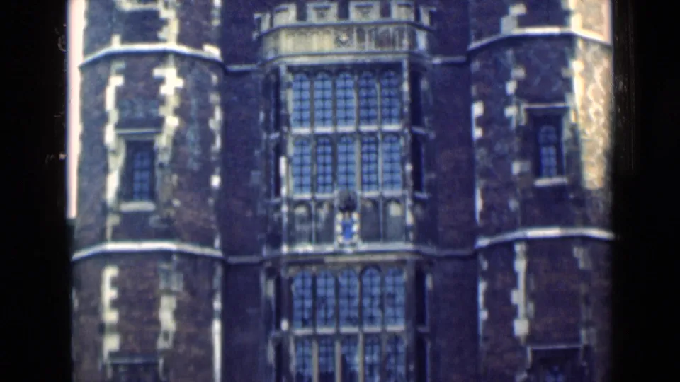 1940s London 8mm frame showing a historic brick facade with stacked leaded windows, a stone balcony, and rounded side towers.