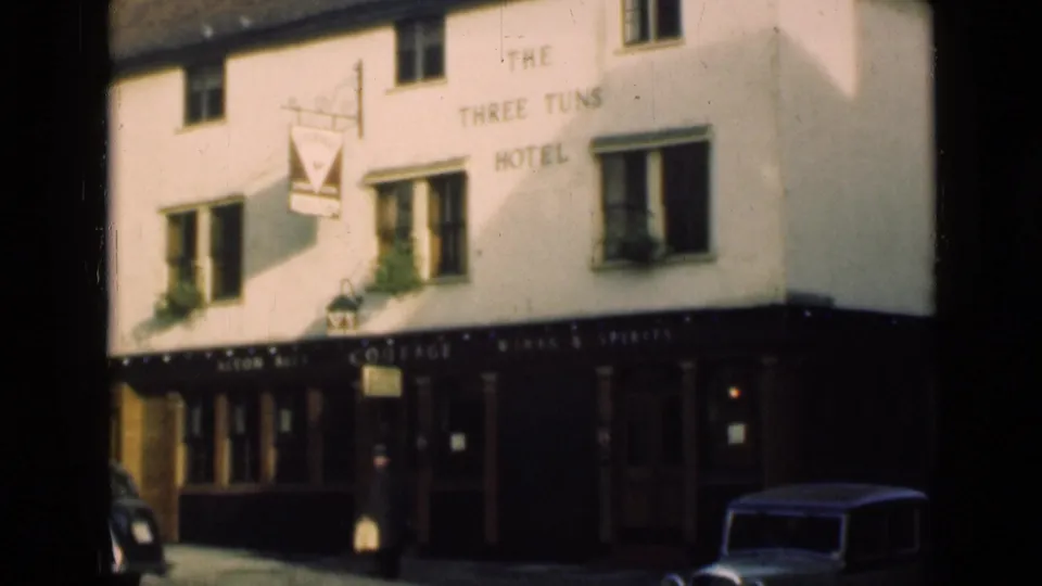 1946 London street frame showing The Three Tuns Hotel frontage, hanging sign, upper windows, a pedestrian, and a parked older car.