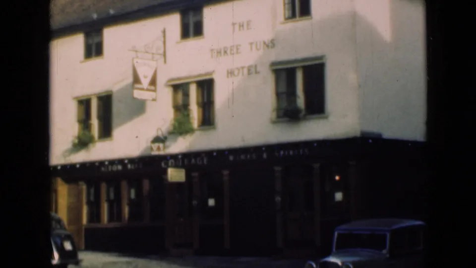 1946 London home-movie frame of The Three Tuns Hotel exterior, with white walls, pub sign, dark trim, and partial classic cars.