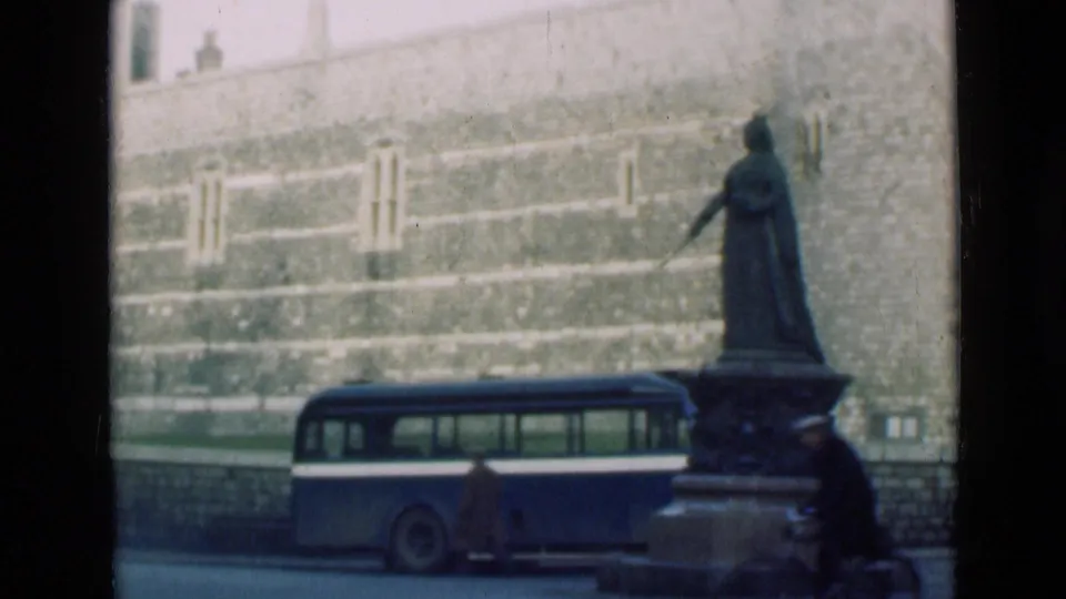 London street view with a statue on a plinth, a bus at the curb, a pedestrian and a large stone wall behind railings.