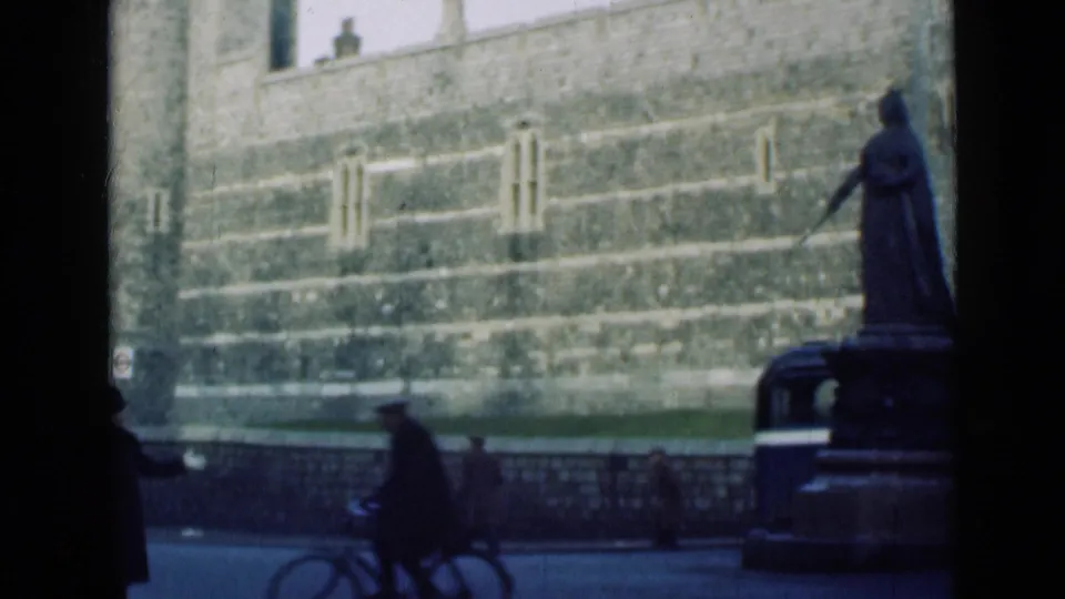 A cyclist and pedestrians move through a London street scene beside a statue, railings and a large historic stone wall.