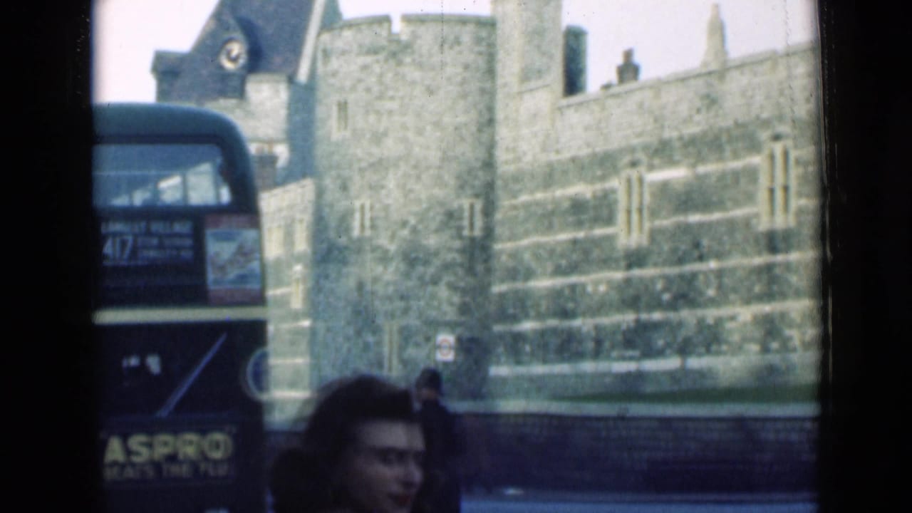 1940s London home-movie frame with a dark double-decker bus, a pedestrian face in the foreground, and a turreted stone building beyond.