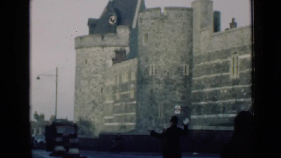 1940s London home-movie frame showing pedestrians in dark coats, a vehicle at the curb, and a turreted stone building beyond.
