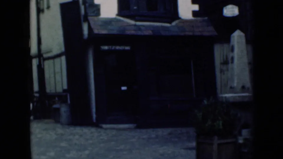 1940s London street-level frame with a shadowed storefront or doorway, stone wall edge, pavement, and visible 8mm frame borders.