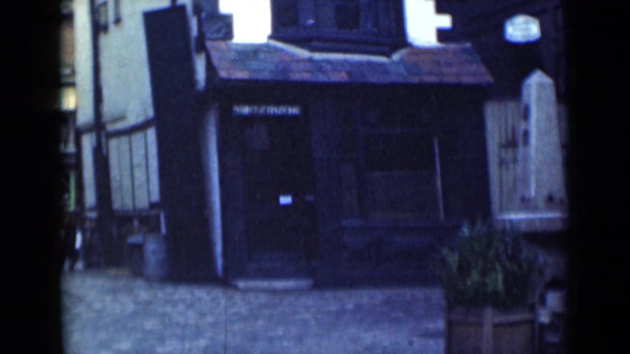 1946 London home-movie frame showing a cobbled street and dark timber-framed building frontage with a small sign and planter.
