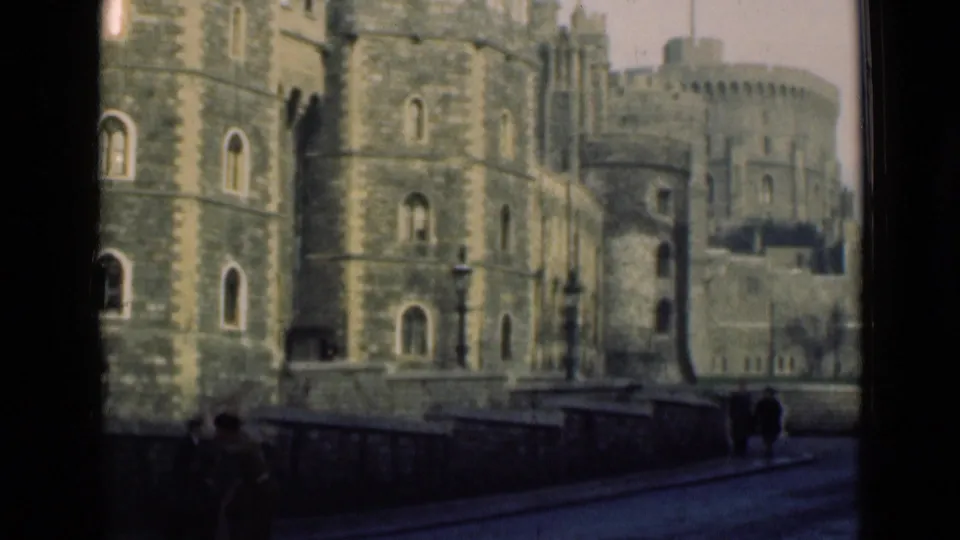 1946 London frame showing castle-like stone masonry, arched windows, a roadside wall, and distant pedestrians.