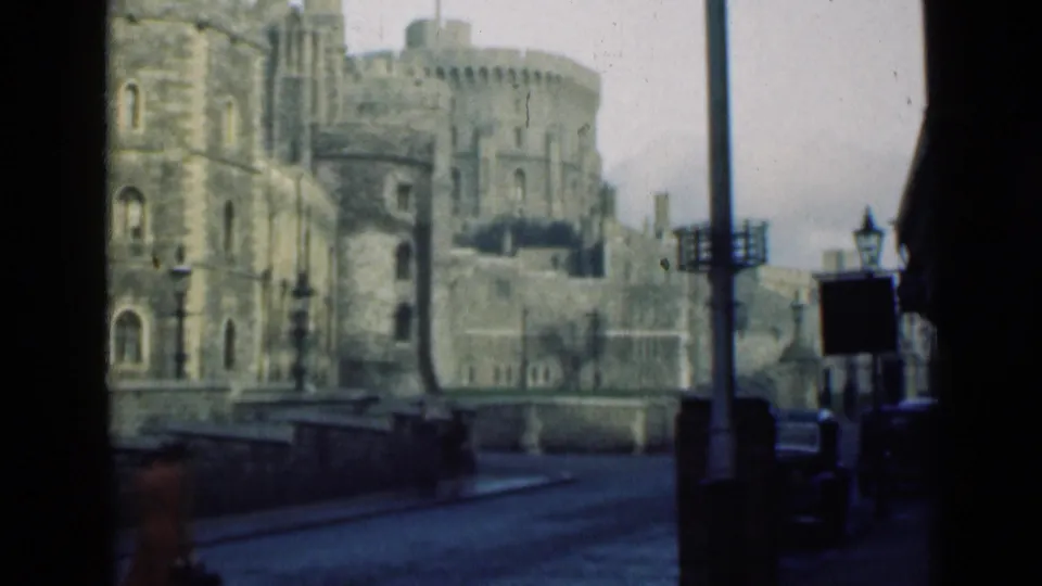 1946 home-movie frame of Windsor Castle from the street, with a blurred passerby shape, lamps, walls, and towers visible.