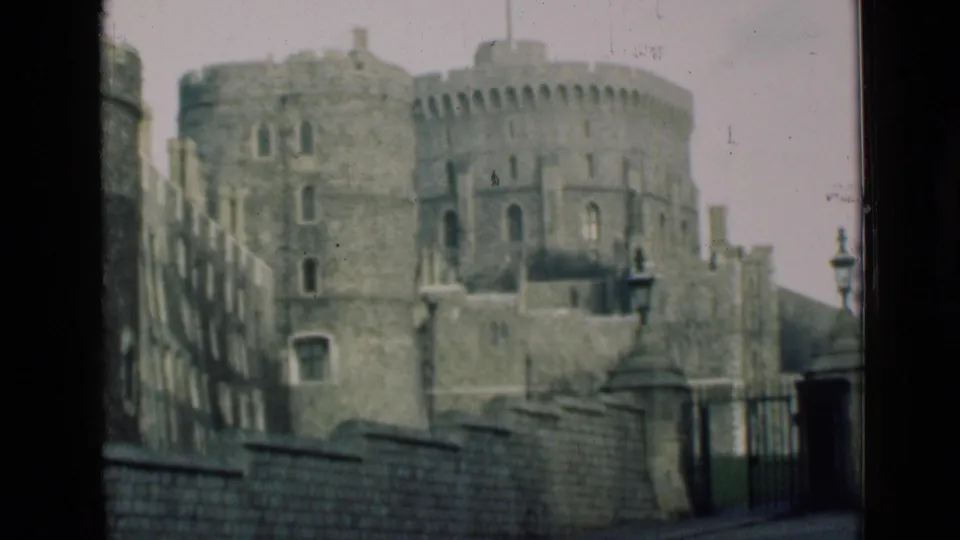 The 1946 home-movie frame shows Windsor Castle's stone towers and battlements beyond a foreground wall, with a gate and lampposts near the right side.