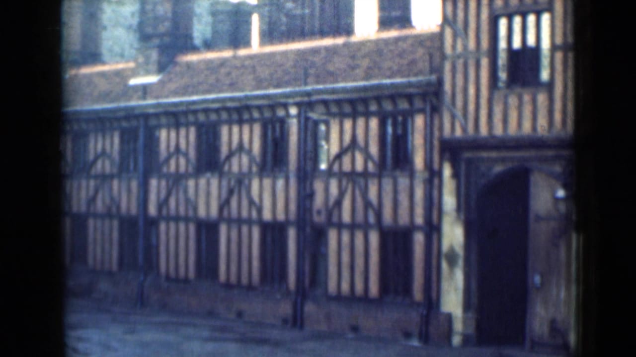 1946 London home-movie frame showing a historic timber-framed facade, dark arched doorway, roof tiles, and pavement.
