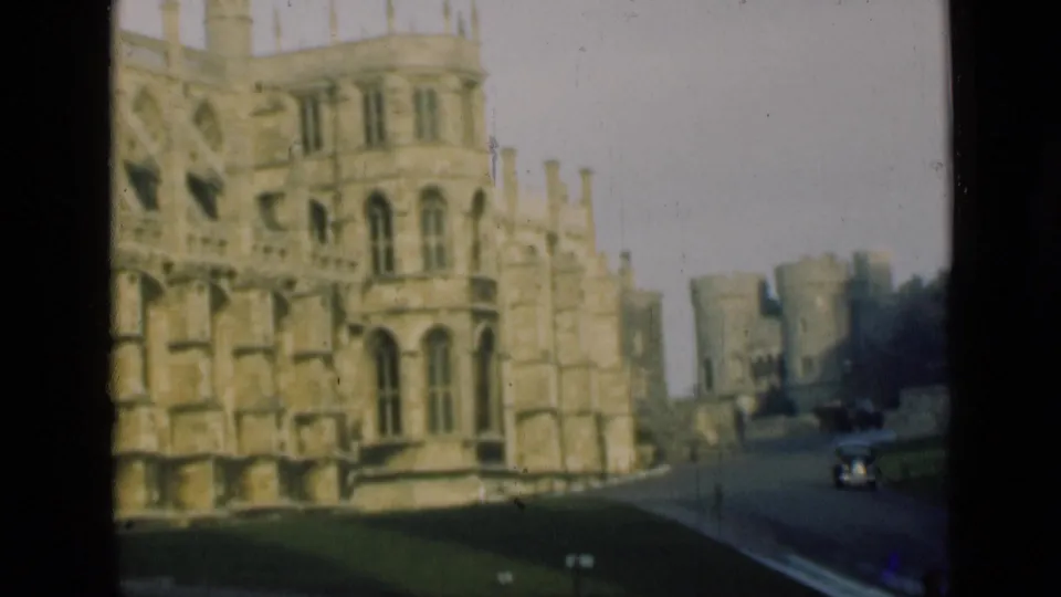 1946 London-area home-movie frame of a Gothic stone castle facade, lawn, and an approaching vintage car on the road.