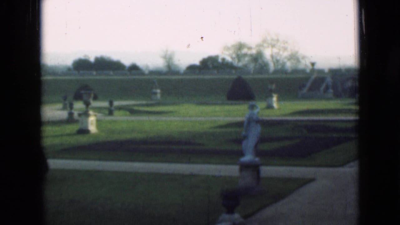 1946 London home-movie frame showing a formal garden through dark window edges, with clipped lawn, pale paths, stone figures, urns, and trees beyond.