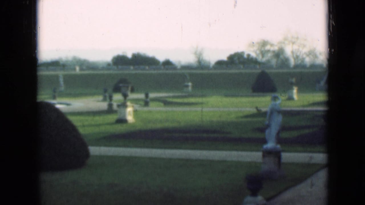 1946 London 8mm frame of a formal garden, with clipped lawn, white garden ornaments, pale paths, distant trees, and a rounded topiary mound.