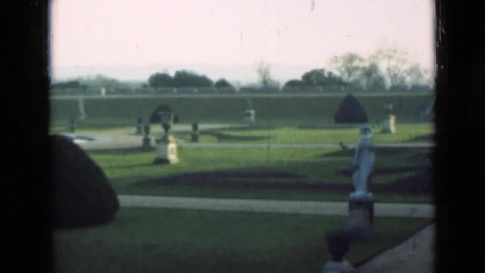 1946 London 8mm frame of a formal garden, with clipped lawn, white garden ornaments, pale paths, distant trees, and a rounded topiary mound.