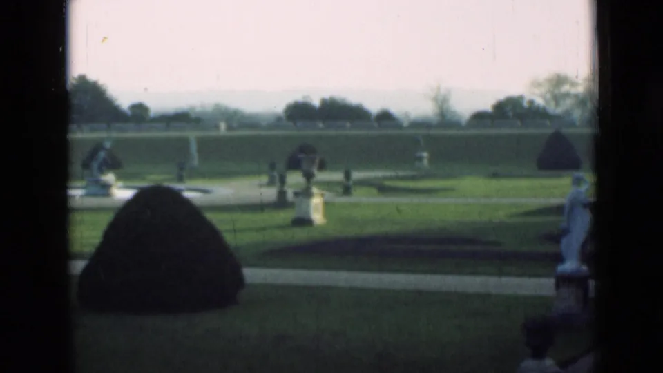 1946 London home-movie frame looking across a formal garden, with a dark topiary mound in front, lawns, white statues, urns, and a pale path.