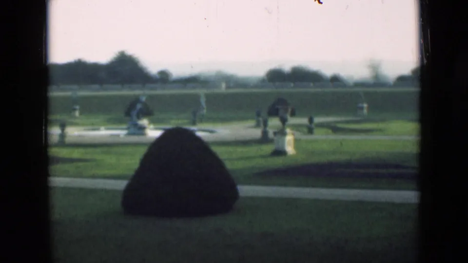1946 London 8mm still of a formal garden with geometric lawns, a rounded topiary mound, white stone garden figures, urns, paths, and distant trees.