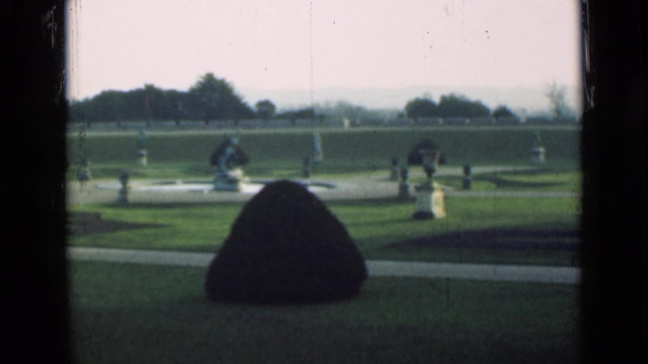 1946 London home-movie view of a formal garden with clipped lawn, conical shrub, classical statues, paths, and distant trees.