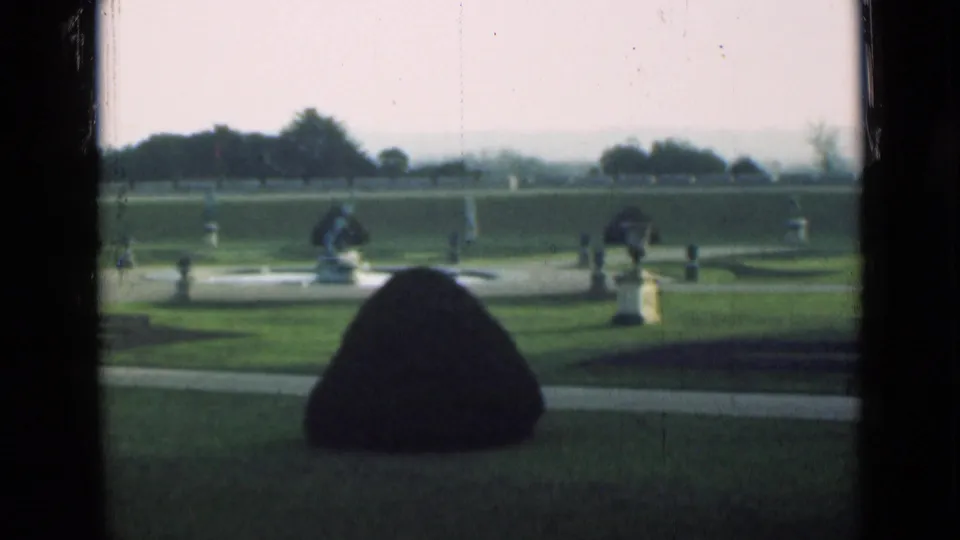 1946 London home-movie view of a formal garden with clipped lawn, conical shrub, classical statues, paths, and distant trees.