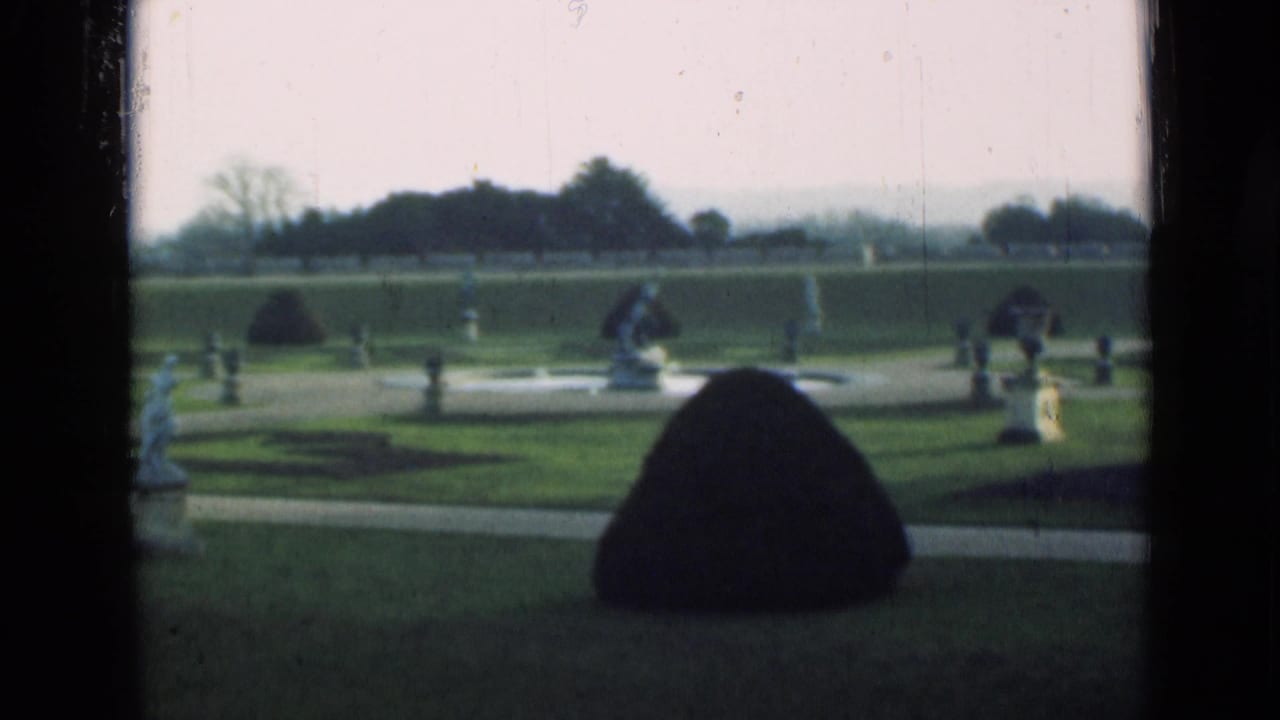 Formal garden lawn with a conical shrub, low paths, statues, and hazy trees in the distance.