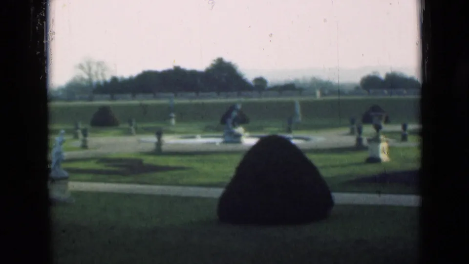 1946 London garden view showing clipped grass, a large shaped shrub, ornamental statues, path lines, and a hazy treeline.