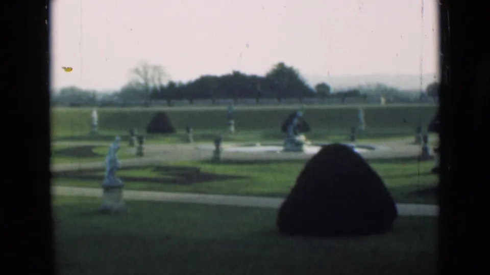 1946 London formal garden shot with a statue at left, shaped shrub at right, intersecting paths, lawns, and distant trees.