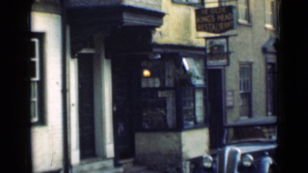 1940s London street view showing a small shopfront, hanging sign, building facades, and part of a classic car at the curb.