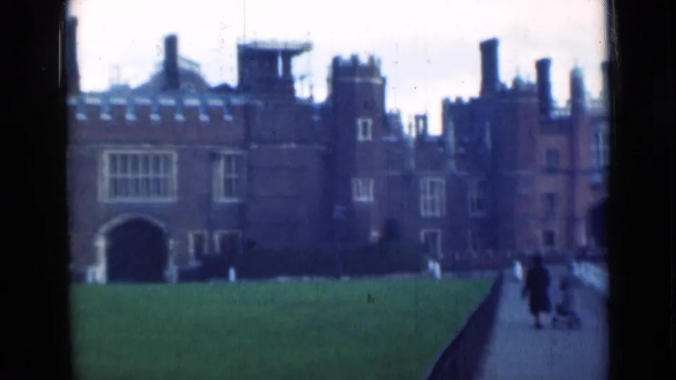London landmark-style exterior with a lawn, arched entrance, chimneys, and distant people walking along a path at the right edge.