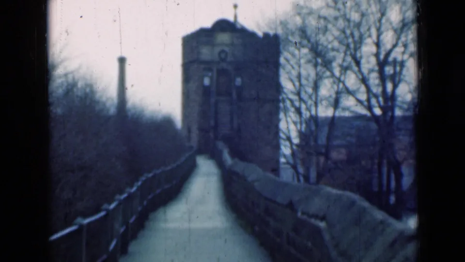 1940s London frame showing a narrow stone walkway leading to a brick tower, with bare trees and railings beside it.
