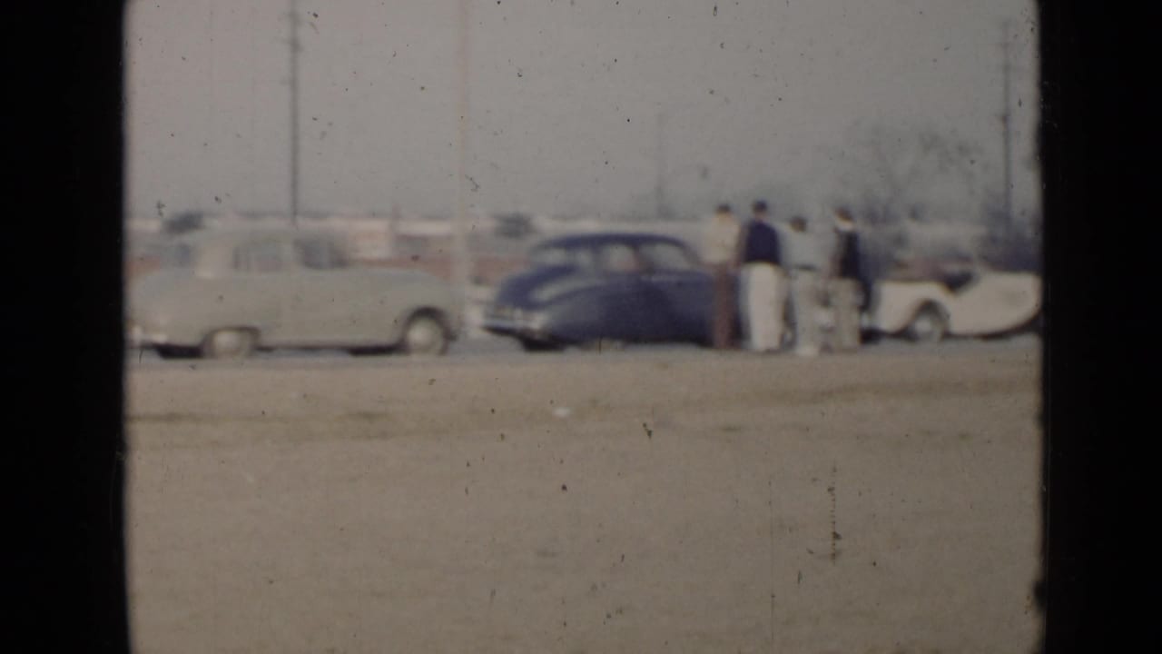 In the San Diego rally footage, several men stand near a light sedan, dark coupe, and pale convertible on a dusty lot.