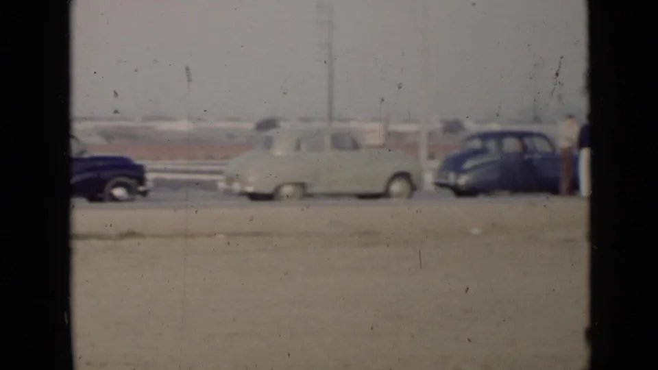 Mid-century cars sit along a dusty roadside in the 1949 San Diego rally footage, with people partly visible near the right edge.