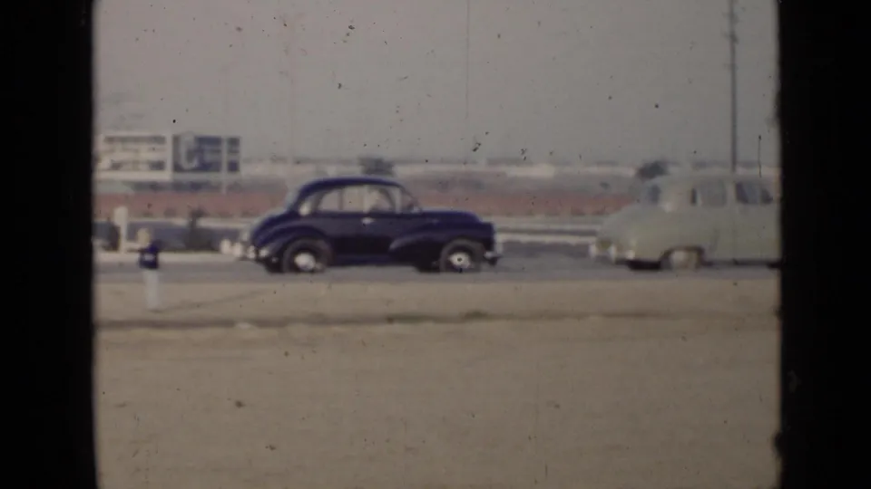 A dark mid-century sedan and a light car are parked beside a dusty roadside in the San Diego rally home movie.