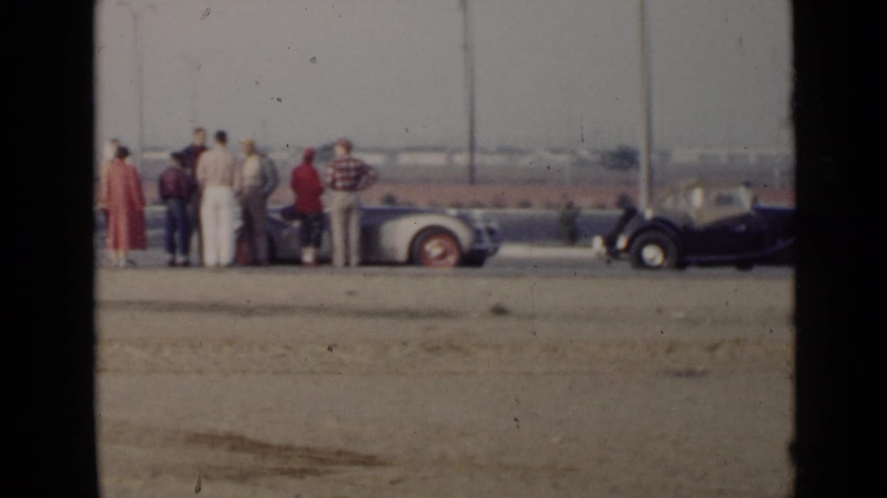 In 1949 San Diego, adults stand near classic convertibles on a dusty lot with open fields and utility poles beyond.