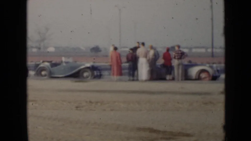 Adults in period clothing stand near parked convertibles in San Diego, with open land and distant structures in the background.