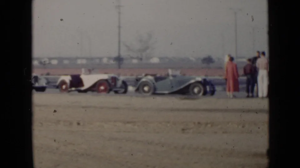 Two classic convertibles and several adults appear on a dusty San Diego lot, filmed in soft color from a distance.