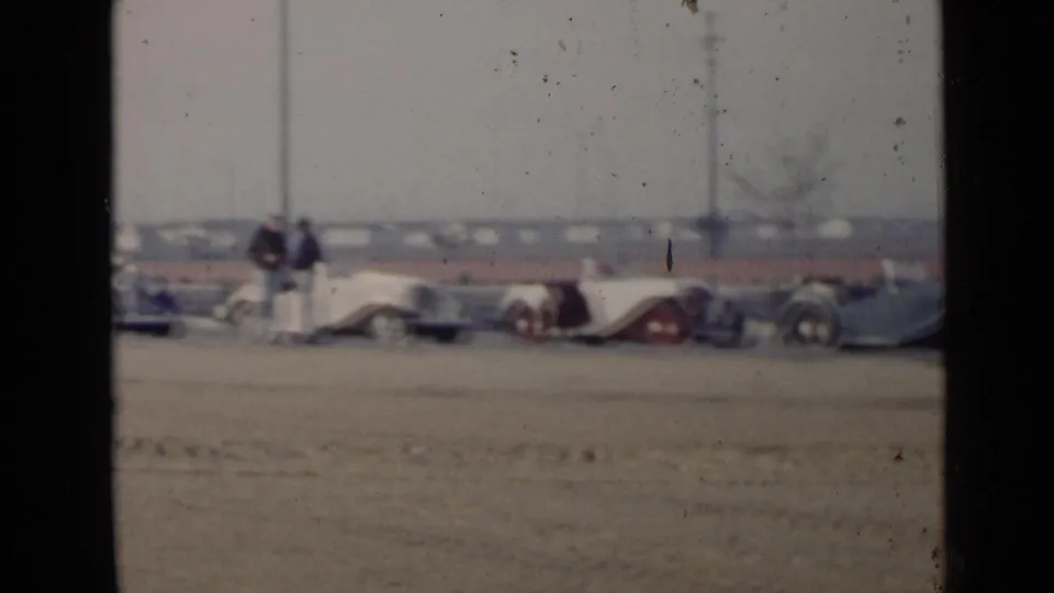 Several classic convertibles are lined up on a dusty San Diego roadside as adults stand between the cars.