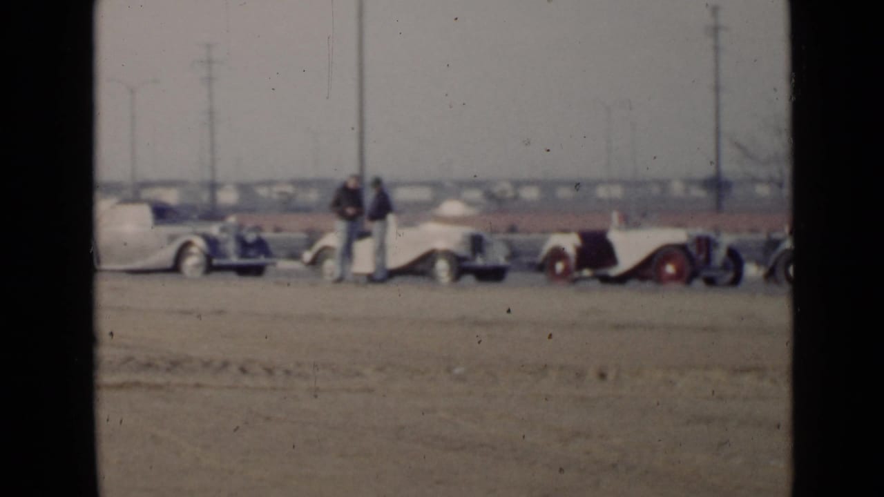 1949 San Diego home-movie frame showing a row of classic cars on a sandy lot with men standing near the vehicles.