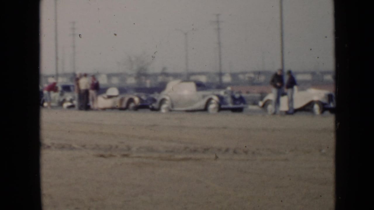 1949 San Diego footage frame with multiple parked 1940s cars and several adults gathered along a sandy roadside lot.