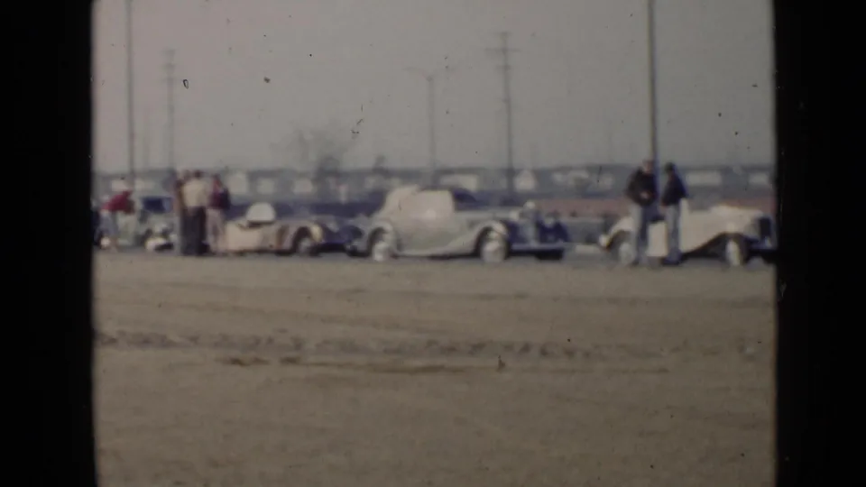 1949 San Diego footage frame with multiple parked 1940s cars and several adults gathered along a sandy roadside lot.