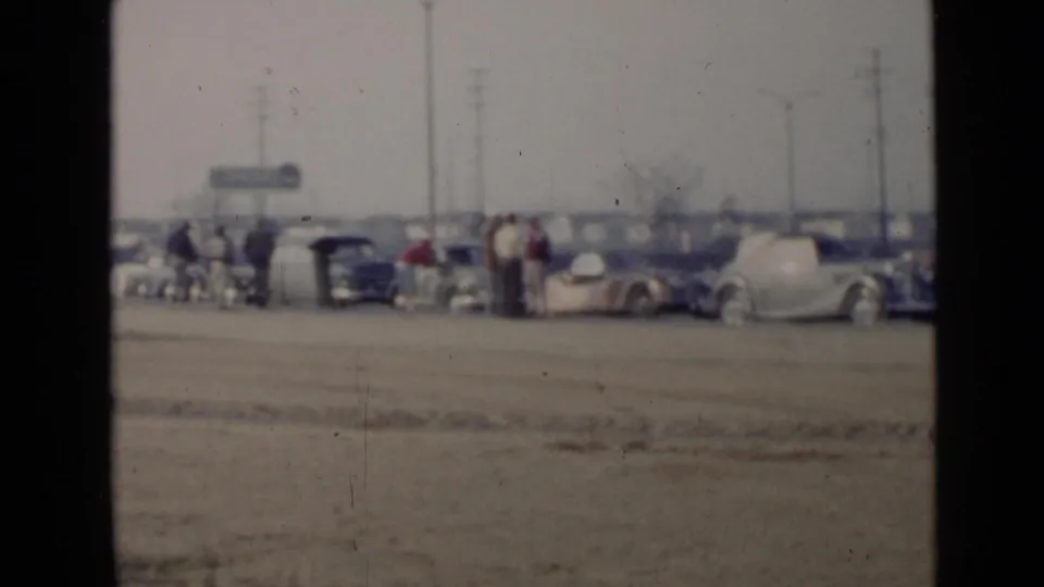 Soft 8mm frame from 1949 San Diego showing adults near classic cars in a broad sandy lot with utility poles behind them.