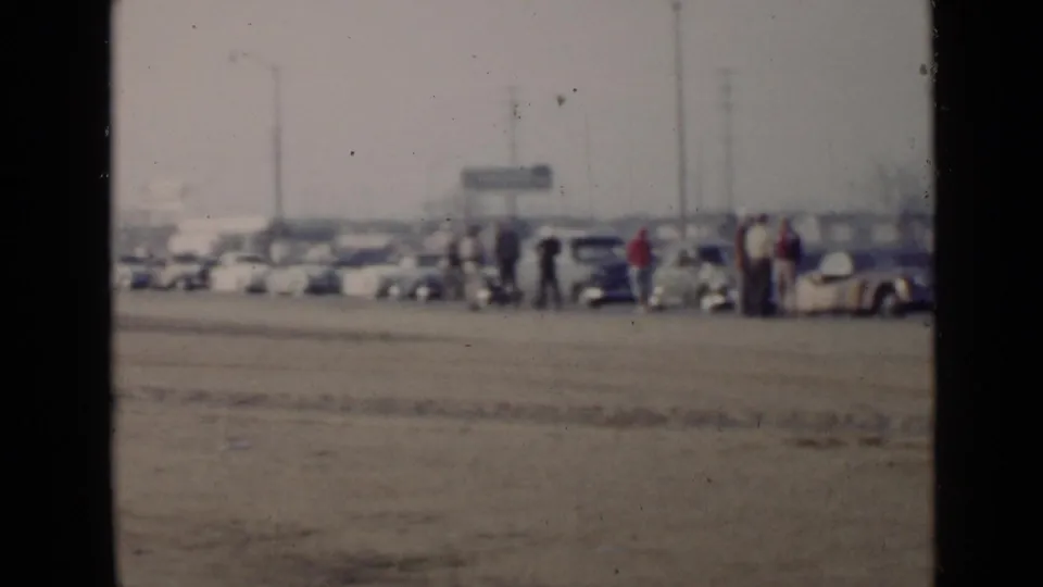 Wide 1949 San Diego home-movie frame with classic cars lined up near adults, utility poles, and distant buildings beyond a sandy lot.