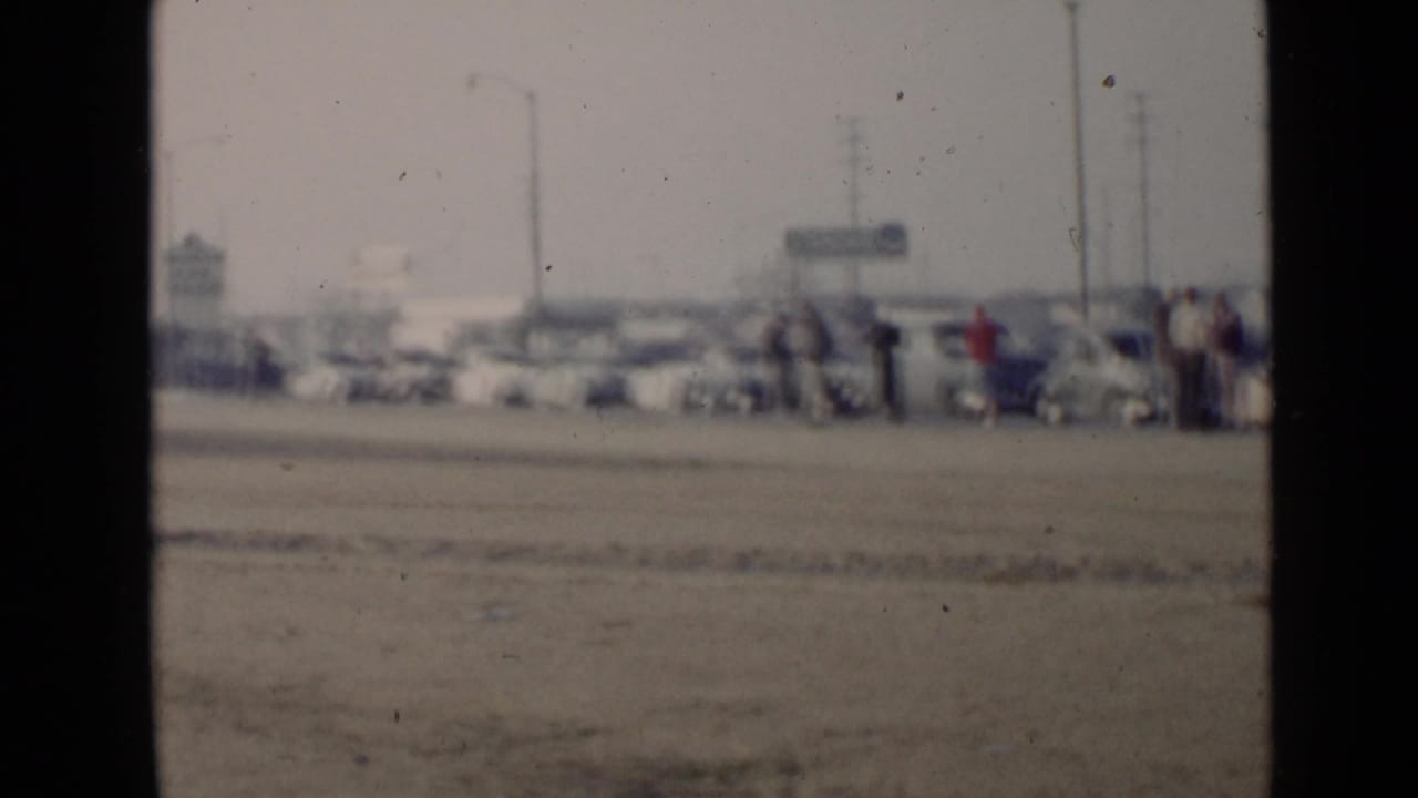 1949 San Diego 8mm frame showing a distant roadside gathering with parked cars and pedestrians in soft focus.