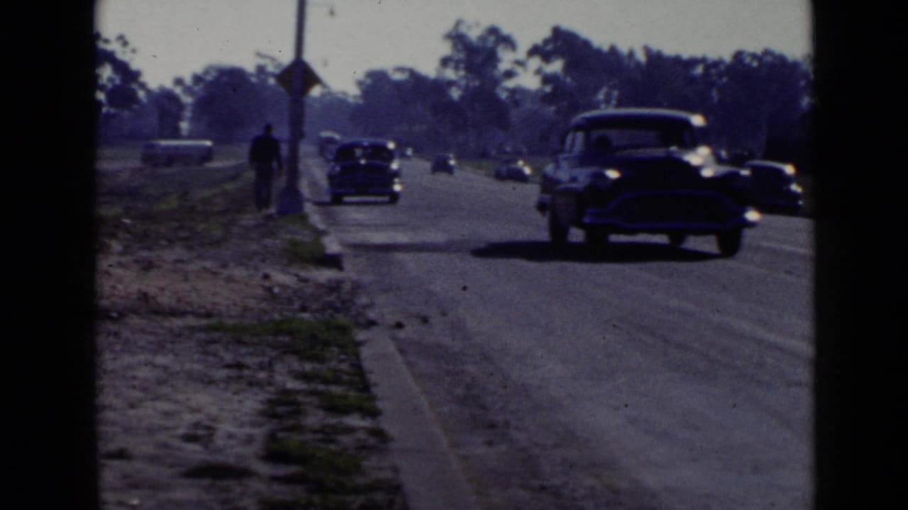1949 San Diego street view with mid-century cars passing and a dark-clothed pedestrian walking beside the curb.